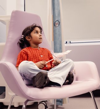 A little girl sits on a chair while in hospital. She holds a wand and looks into the distance.