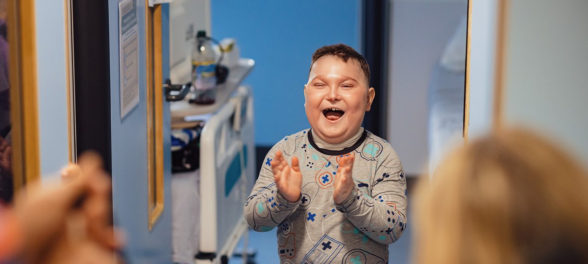 Boy standing in hospital corridor smiling and clapping at entertainment in front of him