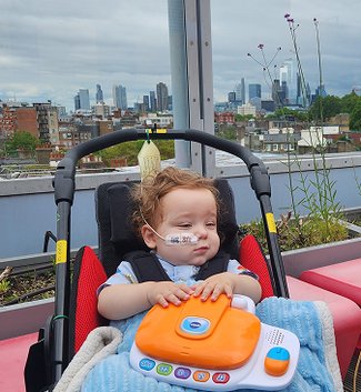 Little boy with feeding tube in buggy on rooftop garden. London skyline visible in the background.