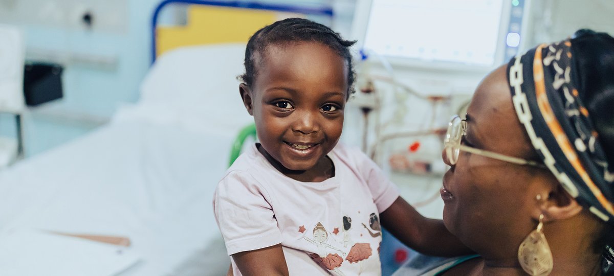 little girl sits on mother's lap, both smiling. They are inside hospital room.