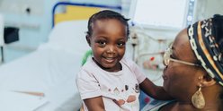 little girl sits on mother's lap, both smiling. They are inside hospital room.