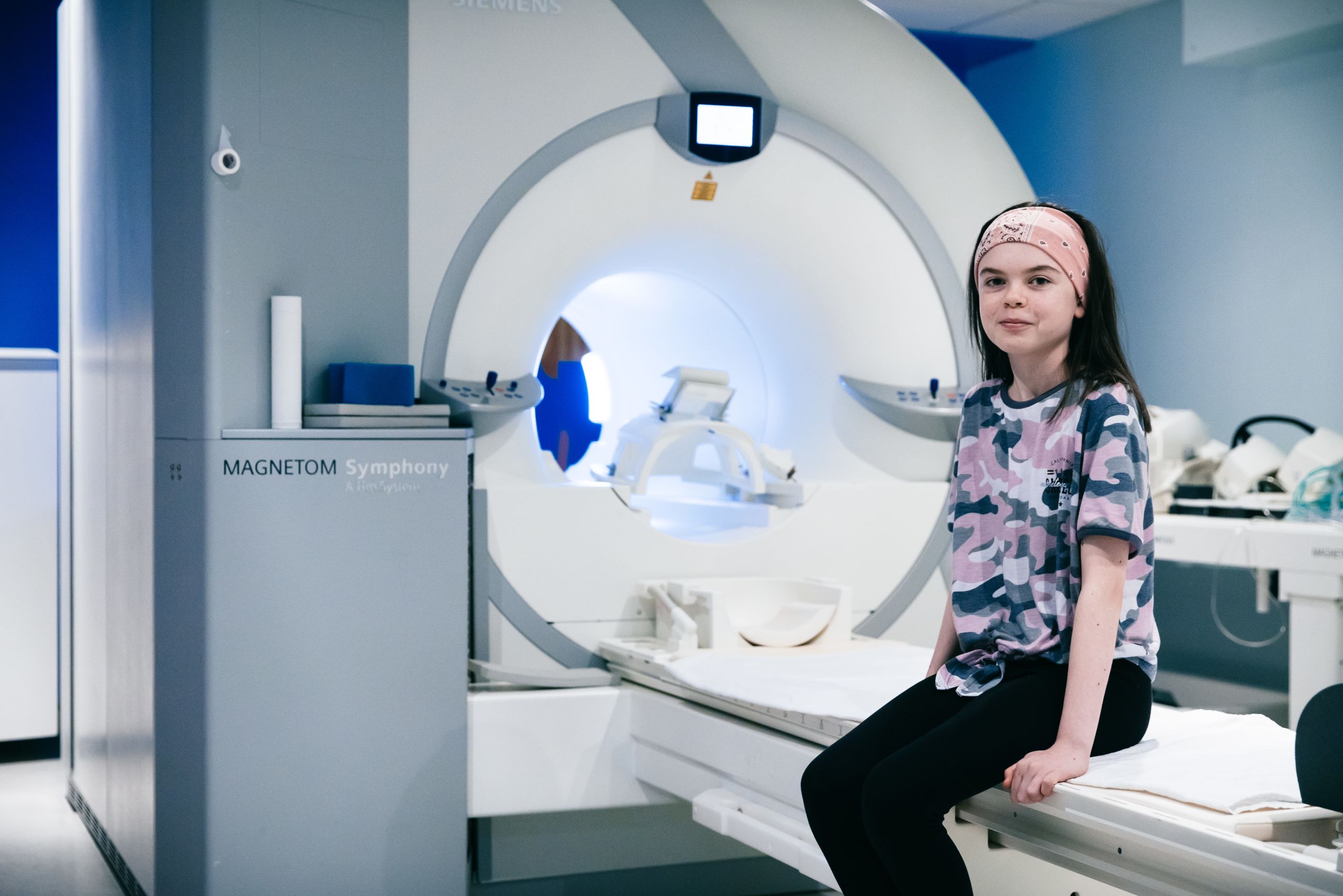 A young girl wearing a pink headband sits on the bed of an MRI scanner.